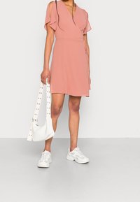 Różowa sukienka kopertowa z krótkimi falbankami na rękawach, sięgająca kostek, zestawiona z białymi trampkami i małą białą torebką z paskiem ozdobionym ćwiekami.