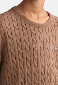 Suéter marrón de punto con cuello redondo y bordes acanalados; presenta un pequeño logo en el pecho. Textura suave, material de peso medio.