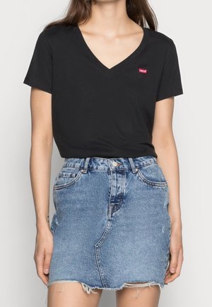 Kobieta ubrana w czarny T-shirt z dekoltem w szpic z małym czerwonym logo oraz niebieską spódnicę dżinsową z wysokim stanem i postrzępionym brzegiem.