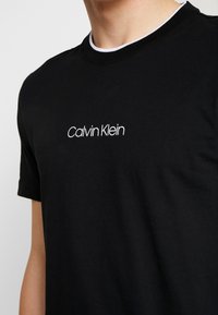 Černé bavlněné tričko s kulatým výstřihem a bílým akcentem. Na přední straně je vyšívaný nápis "Calvin Klein" v bílé barvě.