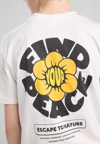 T-shirt bianco in cotone con una grande grafica nera e gialla che recita "TROVA LA TUA PACE" con un design a fiore e il testo in basso "FUGGI NELLA NATURA."