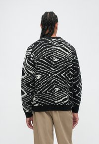 Person mit geflochtenem Haar, die einen schwarz-weißen Pullover mit geometrischem Muster und beigefarbene Hose trägt, steht mit dem Rücken zu einer schlichten Hintergrund.