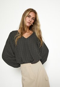 Melnā un baltā rakstainā blouse ar V-veida kakla izgriezumu un br�īviem piedurknēm, savienota ar beigē krāsas svārkiem, kuriem ir sānu kabatas. Mīksta, viegla auduma.