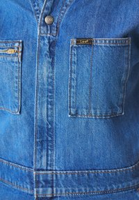 Gros plan de l'avant d'une veste en denim bleu montrant un bouton et deux poches poitrine, dont une avec une étiquette de la marque Lee.