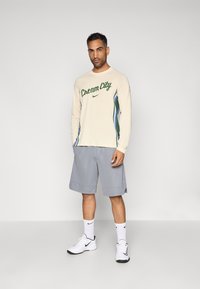 Camisa bege de manga comprida com gráfico "Cream City" em verde, combinada com calções cinzentos, sapatilhas brancas e meias Nike brancas.