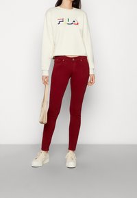 Žena v krémovém svetru FILA, burgundských úzkých kalhotách, krémových sneakers na platformě a držící béžovou kabelku na jednobarevném pozadí.