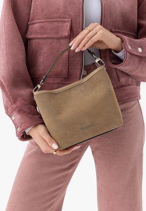 Femeie purtând o jachetă și pantaloni din catifea în nuanță mauve ține o geantă mică din piele întoarsă bej cu mâner negru și accesorii argintii la nivelul taliei.