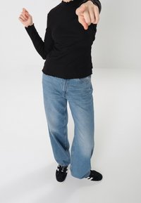 Schwarzes Langarmoberteil mit gewelltem Saum, kombiniert mit weiten, hellblauen Denim-Jeans. Schwarze Sneakers mit weißen Akzenten auf weißem Hintergrund.