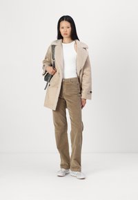 Palton bej din blană sintetică cu guler moale, cămașă albă, pantaloni din corduroy bej, și adidași gri și albaștri, împreună cu o geantă de umăr gri.