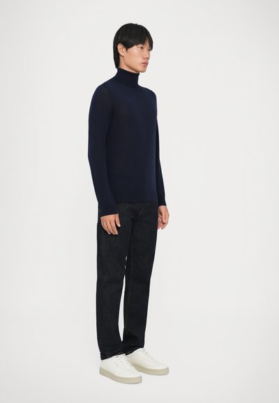 Marineblauer Rollkragenpullover aus weichem Material, kombiniert mit dunklen Jeans und weißen Sneakers, der ein figurnahes Design und klare Linien zeigt.
