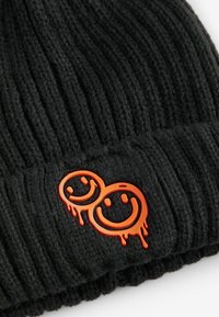 Schwarze gerippte Strickmütze mit zwei orangen lächelnden Gesichtern als Logo-Detail, mit einem tropfenden Farbeffekt am unteren Rand.