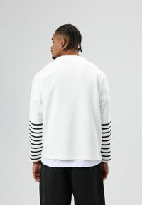 Witte sweatshirt met zwarte horizontale strepen op de mouwen, een ronde hals en een losse pasvorm, passend bij een gelaagde zwarte outfit.