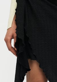 Κοντινό πλάνο ενός χεριού που ακουμπά δίπλα σε μαύρο υφασμάτινο ύφασμα με σατινέ άκρες, σε συνδυασμό με ένα μπεζ μακρυμάνικο ένδυμα.