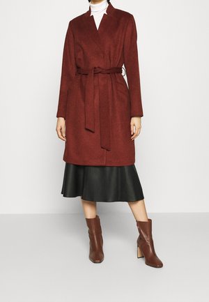 Femme portant un manteau ceinturé couleur rouille sur une jupe midi noire et des bottines marron, debout devant un fond uni.