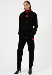 Zwarte hoodie en joggers van zachte stof, met rode logo patches en trekkoorddetails, gecombineerd met zwarte enkellaarsjes.