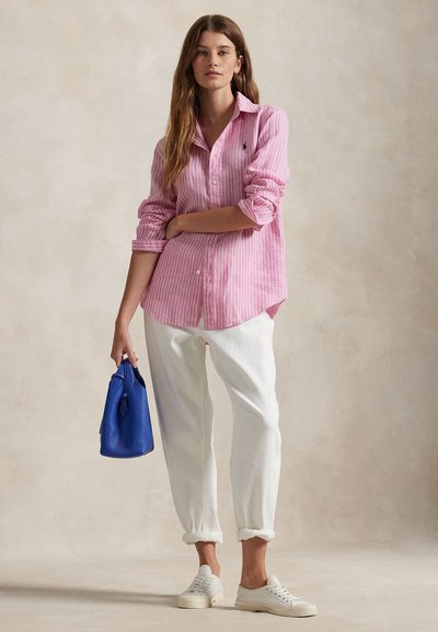 Kvinde iført lyserød, stribet skjorte med knapper, hvide opkrogede bukser, hvide sneakers, holder blå håndtaske, står foran en ensfarvet baggrund.