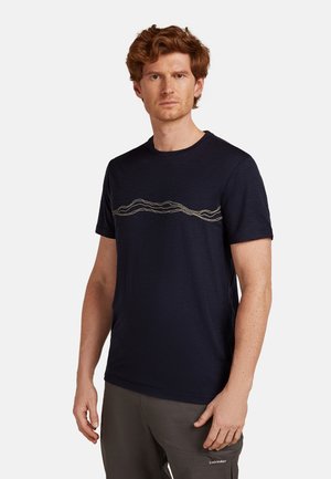 Mann mit roten Haaren, der ein navy-blaues T-Shirt mit einem beigen Wellenmuster über der Brust und grauen Hosen mit einem Icebreaker-Logo trägt.