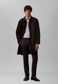 Manteau noir léger avec fermeture à boutons, le modèle porte une chemise rayée et un pantalon foncé. Tissu lisse, coupe longueur genou.