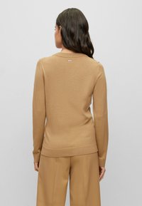 Beżowy sweter dzianinowy z długimi rękawami, okrągłym dekoltem i małym logo na plecach. W zestawie ze współczesnymi szerokimi spodniami w kolorze beżowym.