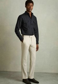 Cămășă neagră cu mânecă lungă, cu două buzunare frontale, asociată cu pantaloni de costum bej și mocasini închiși la culoare, pe un fundal neutru.