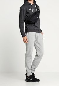 Person som bär en mörkgrå Champion-hoodie, ljusgrå sweatpants, svarta Nike Air Max-sneakers och en svart axelväska.