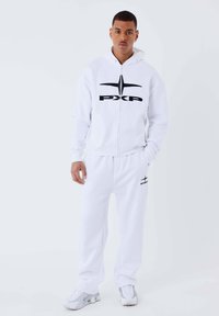Sudadera blanca con cremallera y logo negro "PXP" acompañado de pantalones de chándal a juego. Hecha de tejido suave, presenta un ajuste holgado y puños elásticos. Zapatillas grises.