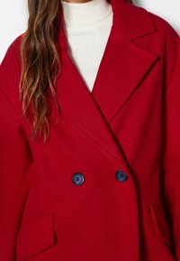 Abrigo rojo de doble botonadura hecho de una mezcla de lana, con cuello de muesca, dos bolsillos frontales y botones azul marino, con una textura suave y lisa.