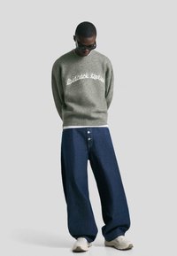 Person, der Sonnenbrillen trägt, einen grauen Pullover mit dem Text "laidback stories", weite dunkelblaue Jeans und weiße Sneaker vor einem einfarbigen Hintergrund.