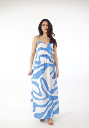 Kobieta o długich włosach, ubrana w niebiesko-białą, abstrakcyjną, bezrękawnikową suknię maxi oraz sandały z przezroczystymi paskami, stojąca na białym tle.