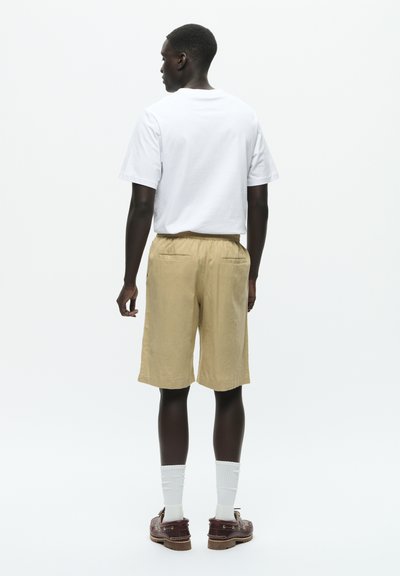 Homme portant un t-shirt blanc, un short beige, des chaussettes blanches et des chaussures bateau marron, debout de dos sur un fond blanc uni.