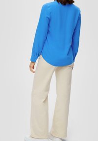 Chemise à manches longues bleu vif avec des poignets boutonnés, assortie à un pantalon large beige clair et des baskets blanches. Texture de tissu lisse.