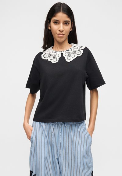 Femme portant un haut noir à manches courtes avec un col en dentelle blanche et un pantalon rayé bleu, les mains dans les poches, sur un fond uni.