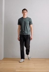 Grå t-skjorte med rund hals, svarte denimjeans og hvite sneakers med grønne detaljer, står på tregulv mot en lys vegg.