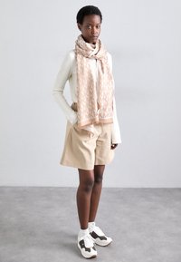 Vrouw die binnenshuis staat, gekleed in beige korte broek, witte trui, een designer sjaal in bruin en beige, witte sokken en witte sneakers met bruine accenten.