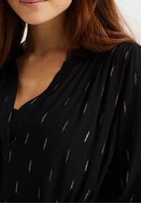 Černá blůza s výstřihem do V, lehká, texturovaná tkanina s metalickými zlatými a stříbrnými listovými vzory po celém povrchu.