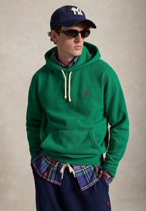 Homme portant une casquette bleu foncé, des lunettes de soleil noires, un sweat à capuche vert par-dessus une chemise à carreaux, un pantalon bleu marine et une montre-bracelet, debout les mains dans les poches.