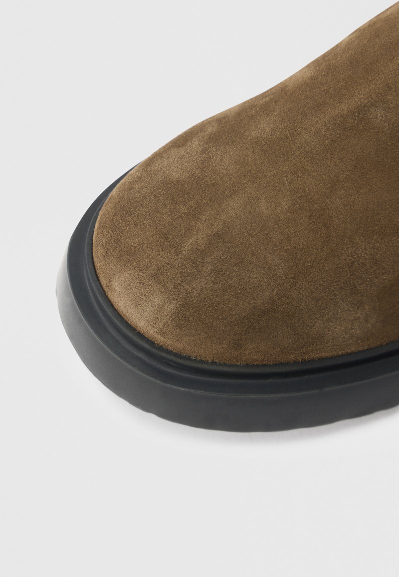 Stivale in camoscio marrone con punta arrotondata e suola in gomma nera a contrasto. La texture è morbida e il design è minimalista senza accenti visibili.