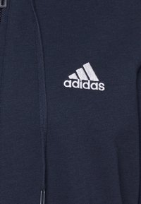 Tissu bleu marine avec logo Adidas blanc brodé sur le côté droit de la poitrine, présentant trois bandes diagonales et le nom de la marque.