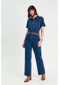 Salopette di denim con maniche corte, due tasche sul petto e vita con cintura. Colore blu scuro con cuciture in contrasto. Taglio a gamba ampia. Stivaletti marroni alla caviglia.