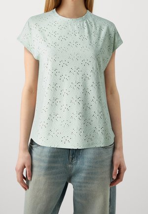 Frau trägt eine hellgrüne Kurzarmbluse mit floralem Lochmuster und ausgewaschene blaue Jeans vor einem schlichten Hintergrund.