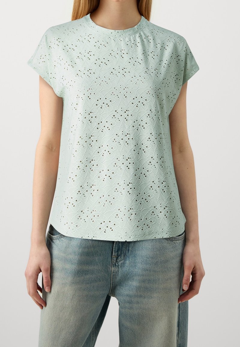 Femme portant un chemisier vert clair à manches courtes avec un motif floral ajouré et un jean bleu délavé sur un fond uni.