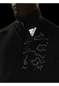 Homme portant un haut Adidas noir zippé avec logo réfléchissant et motif abstrait en points sur la poitrine, visible du cou jusqu'au milieu du torse.