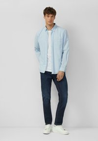 Chemise bleu clair à boutons portée sur un t-shirt blanc, assortie à un jean bleu foncé et des baskets blanches. Dotée d'une poche poitrine et de manches longues.