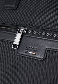 Sac en textile noir avec une fermeture éclair lisse et un tirette métallique. Logo de la marque affiché en cuir noir avec des points d'accent colorés.