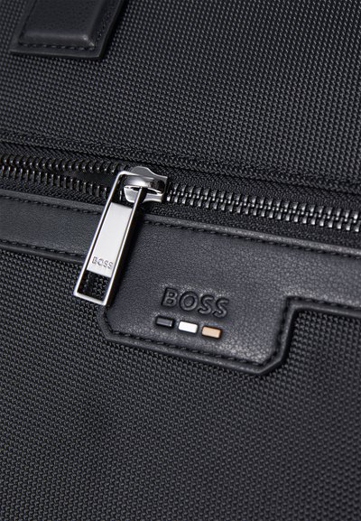 Sac en textile noir avec une fermeture éclair lisse et un tirette métallique. Logo de la marque affiché en cuir noir avec des points d'accent colorés.