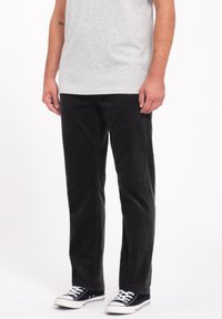 Pantalons en velours côtelé noir avec une coupe droite, présentant des rainures subtiles, associés à des baskets en toile noir et blanc.
