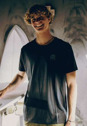 Jeune homme aux cheveux bouclés souriant, portant un t-shirt noir avec un petit logo, debout près d'une planche de surf blanche dans une lumière tamisée.