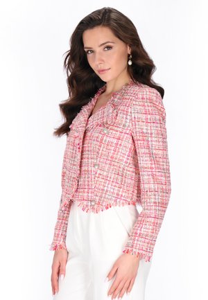 Donna con capelli castani lunghi che indossa orecchini di perla e una giacca in tweed rosa e bianca dalla texture accentuata sopra pantaloni bianchi, posando davanti a uno sfondo bianco.