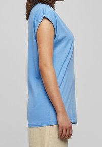 Person i en ljusblå kortärmad skjorta och beiga byxor som står sidan emot en enkel grå bakgrund.