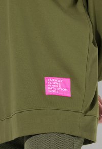 Sweatshirt vert olive en tissu doux, avec un patch rectangulaire rose portant le texte "L'ÉNERGIE FLU D'OÙ L'INTENTION VA" sur le bas.